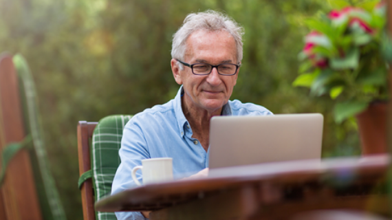 Älterer Herr sitzt mit einem Laptop an einem Gartentisch