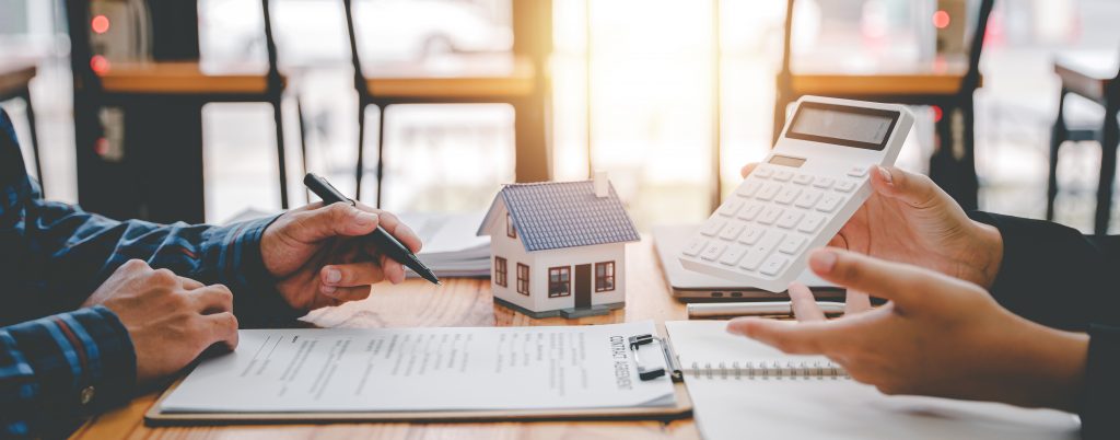 Zwei Personen sitzen an einem Tisch mit Taschenrechner, Miniaturhaus und Unterlagen zur Immobilienfinanzierung.