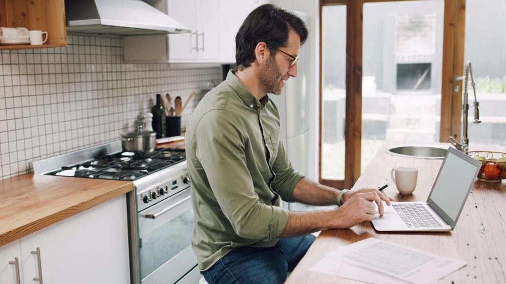 Mann sitzt mit Laptop in der Küche und arbeitet konzentriert.