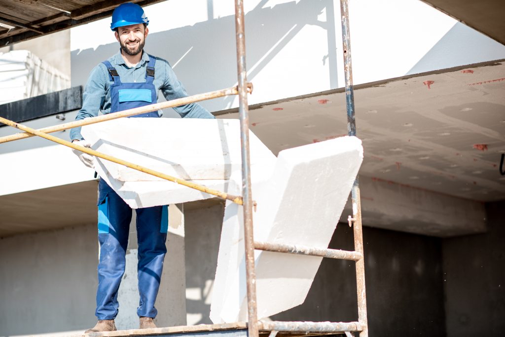 Bauarbeiter auf einem Gerüst hält Dämmmaterial in der Hand vor der Fassade eines Hauses.