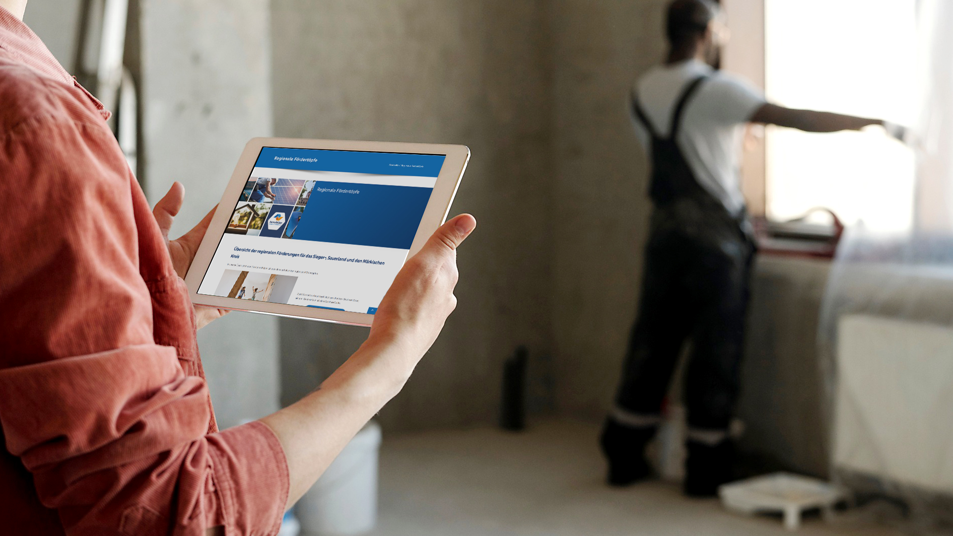 Eine Person hält ein Tablet mit einer Mess- oder Kalibrierungsanzeige in einem renovierungsbedürftigen Raum, während im Hintergrund eine weitere Person an einem Fenster arbeitet. Die Szene zeigt eine Baustellen- oder Modernisierungssituation in einem Innenraum.