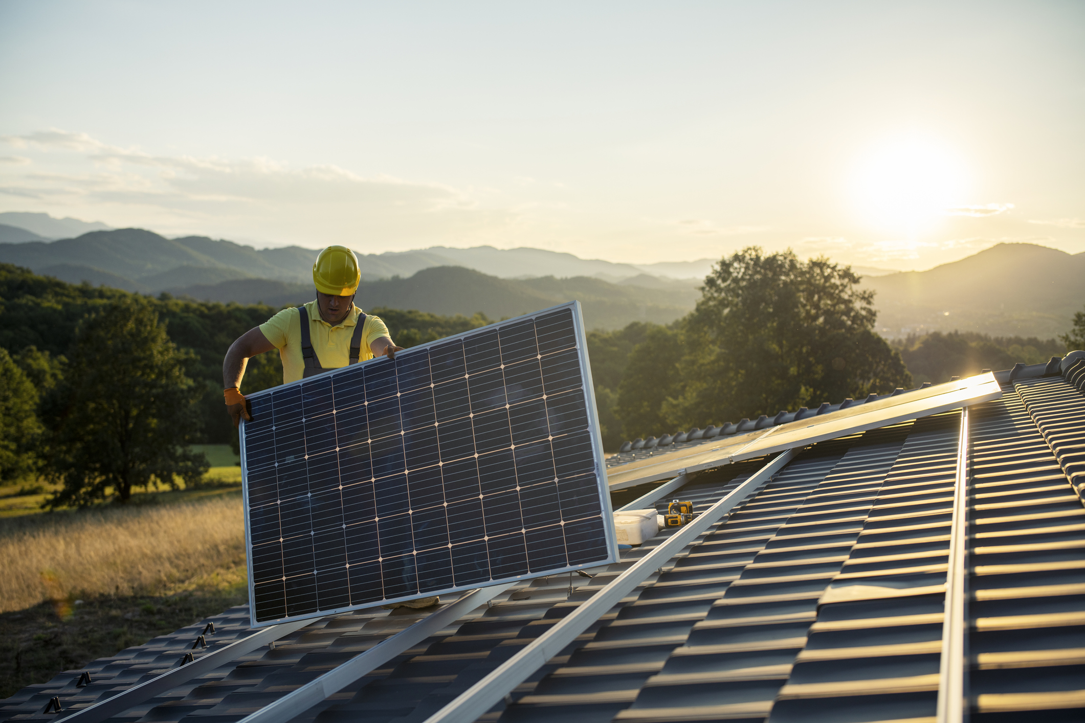 Mann mit Bauhelm auf dem Dach trägt Solarplatte in der Hand.